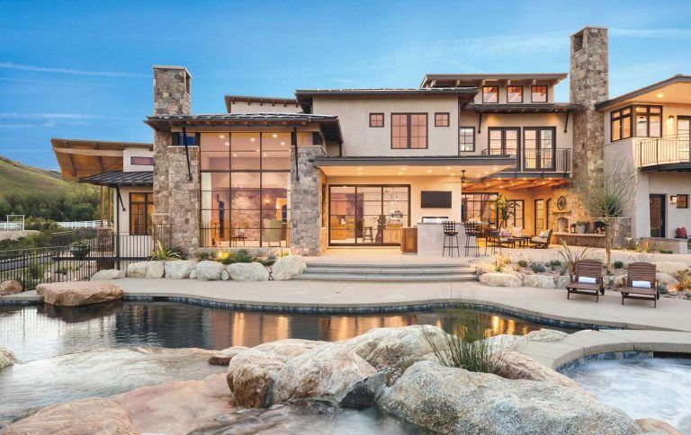 Pool view of large home with floor to ceiling windows, patio furniture, and water features.
