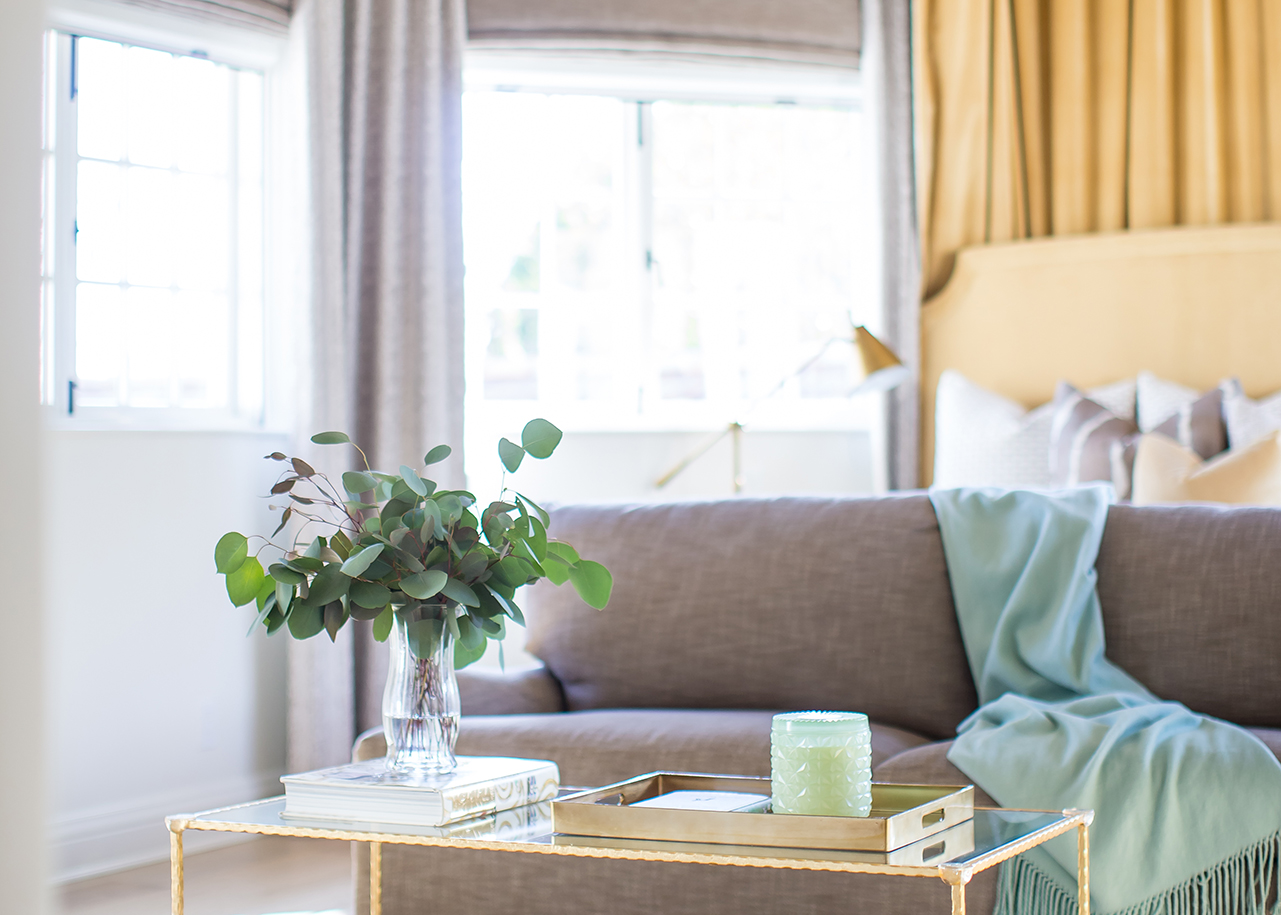 French Normandy-style primary bedroom detail with large bed, draped yellow, grey and teal fabric, gold accents, and eucalyptus in glass vase