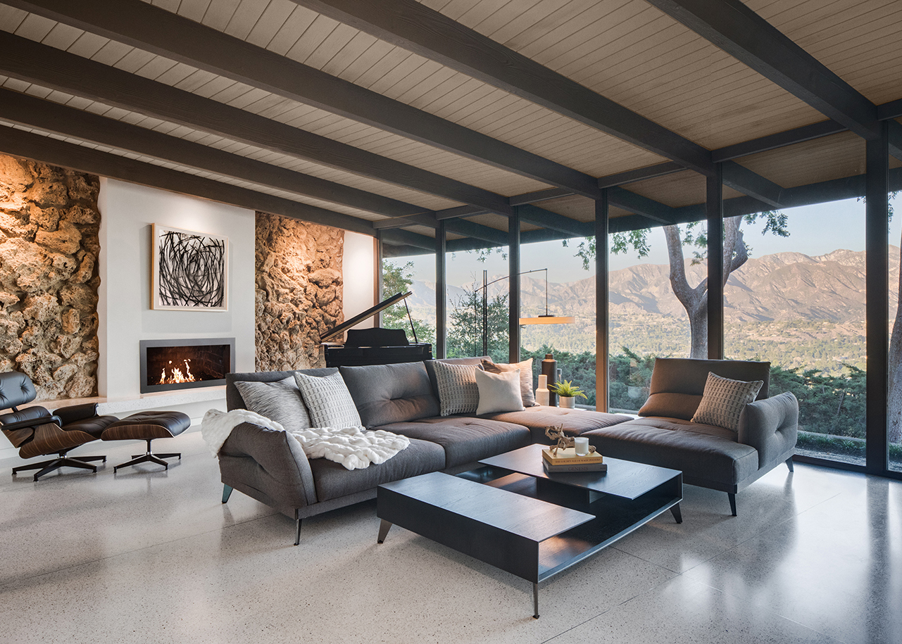Remodeled living room with a sleek fireplace, lava rock wall, Mid-Century Modern furniture, and a wall of windows with views of the San Gabriel mountain range