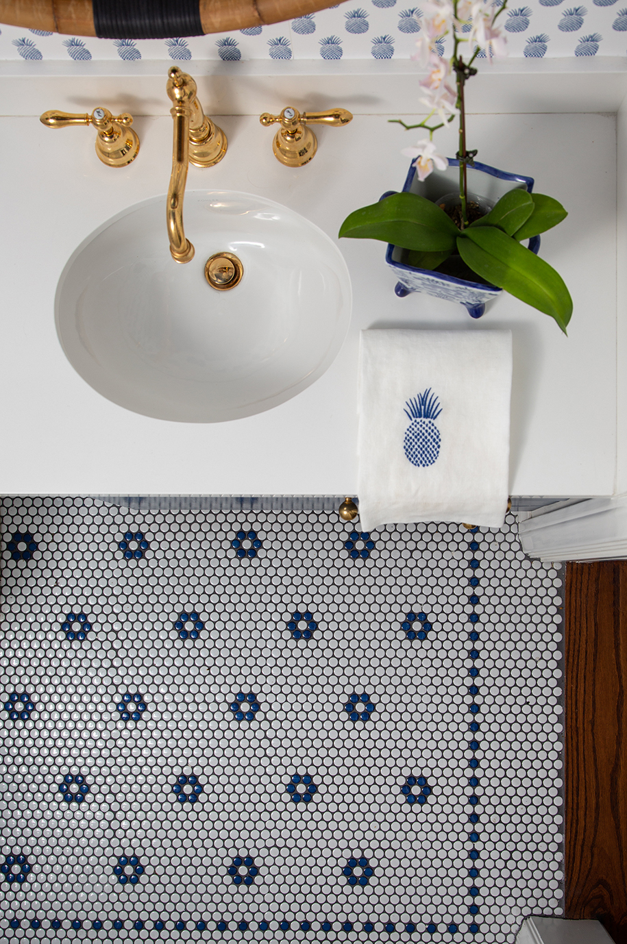 Top down view of white and blue beachy powder bathroom with gold sink, pineapple pattern hand towel, and penny round floor tile with floral motif