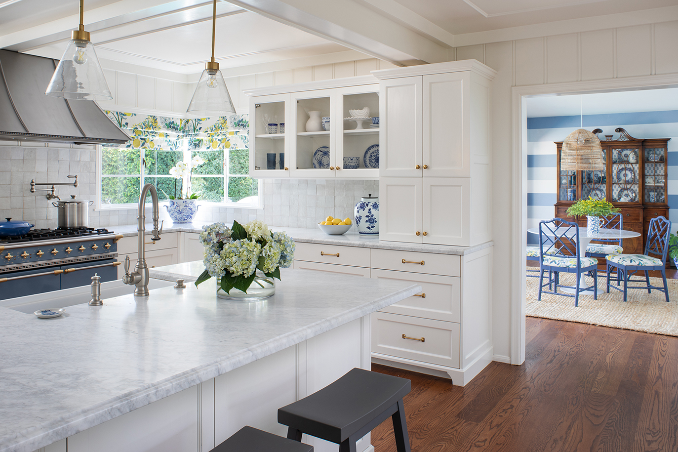 Cape Cod-inspired kitchen remodel with spacious central peninsula with bar seating, white cabinetry, blue Lacanche range, aged brass hardware, and formal dining room with blue striped wallpaper and basket pendant light seen beyond