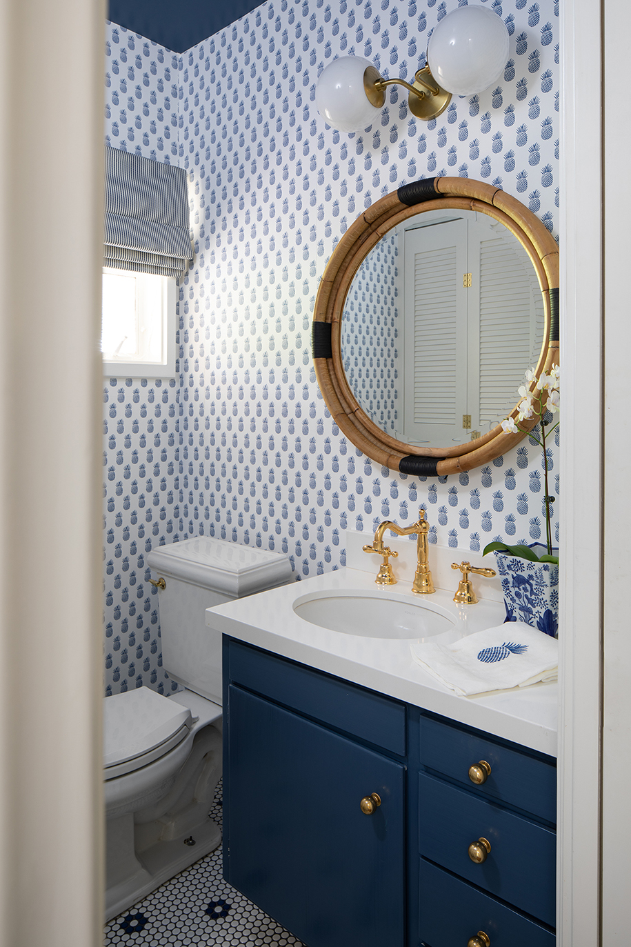 Coastal-inspired powder bathroom remodel with navy blue painted cabinetry, pineapple patterned wallpaper, rattan circular mirror, and penny-round floor tile