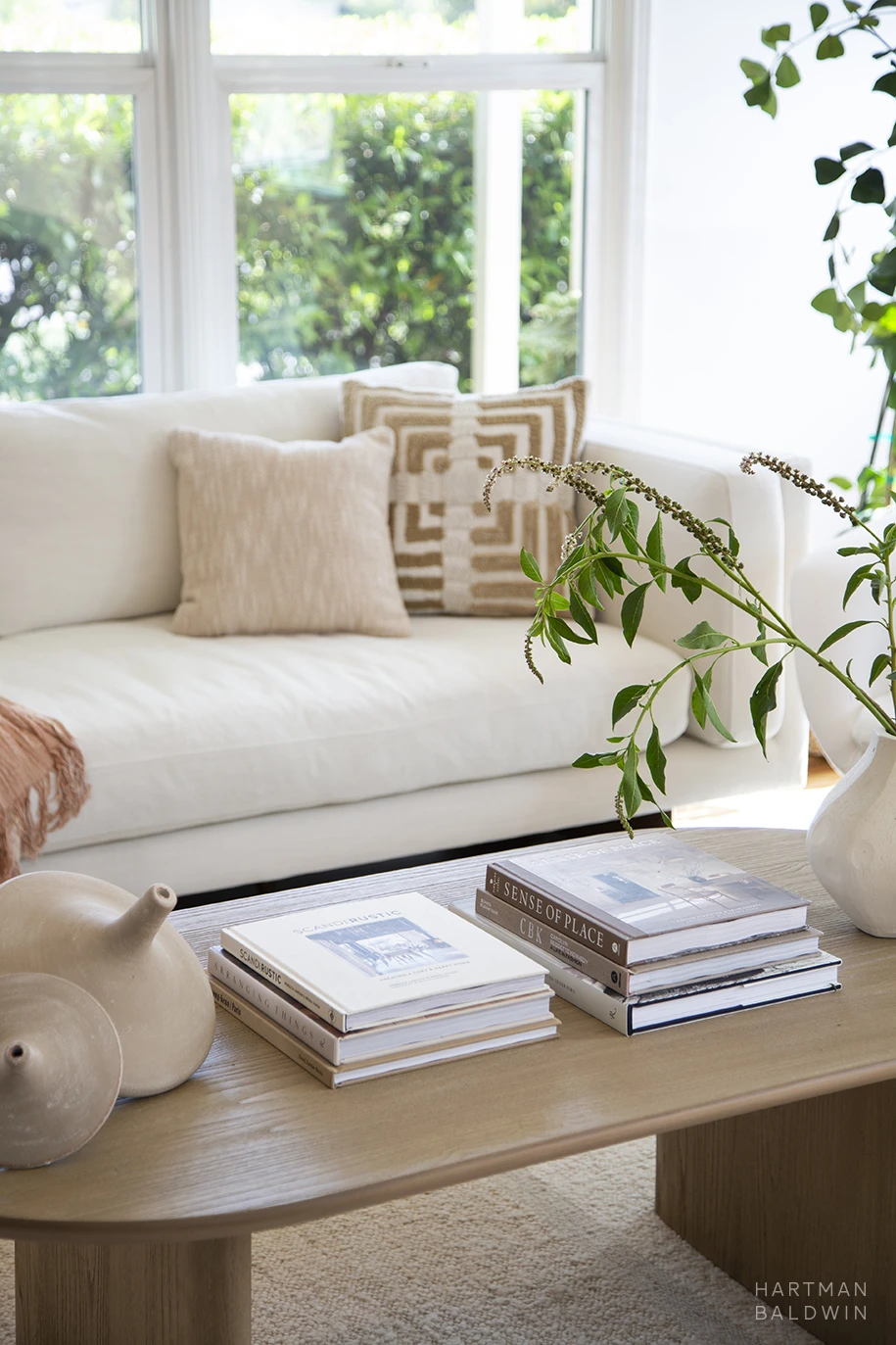 Bright and contemporary remodeled living room with white couch, neutral pillows, and wooden coffee table with interior design photo books, wood accessories, and foliage in white vase