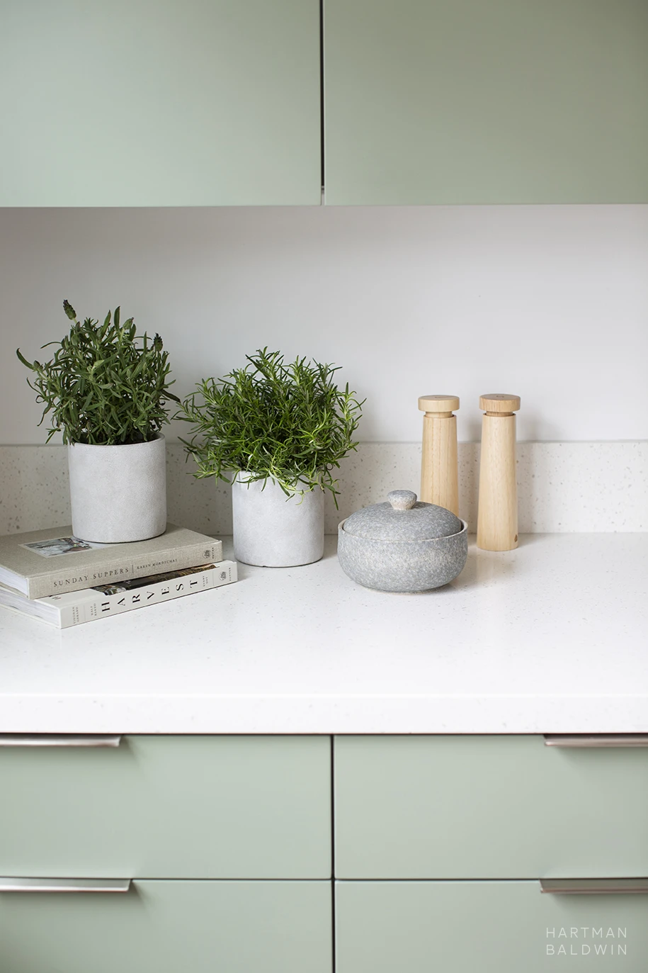 Kitchen accessories, cookbooks, and potted herbs on white quartz countertop surrounded by sage green slab-style cabinetry