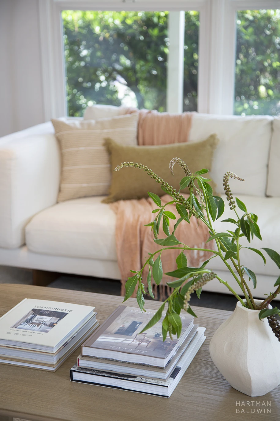 Bright and contemporary remodeled living room with white couch, neutral pillows, pink throw blanket, and wood coffee table with interior design photo books and foliage in white vase
