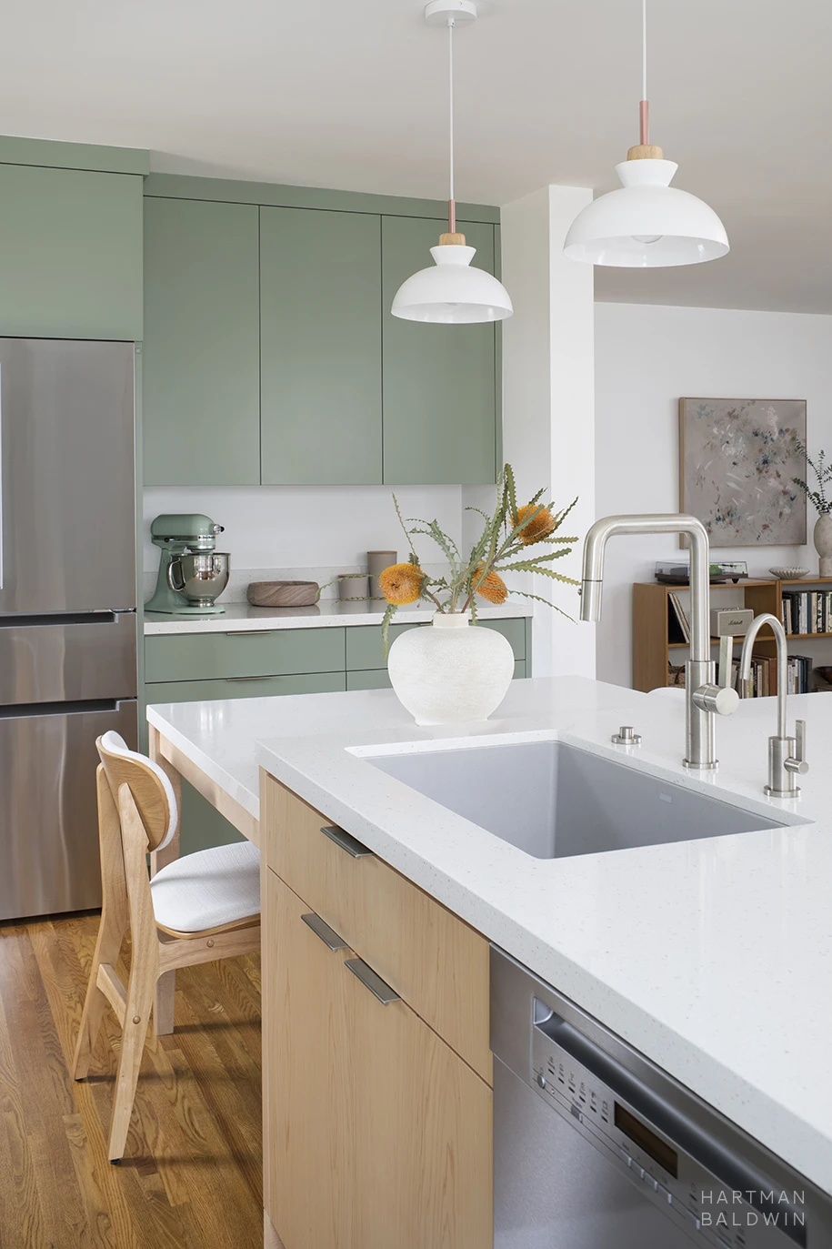 Contemporary Scandinavian minimalist style kitchen remodel with sage green cabinetry, sculptural pendant lights, and a central island with a built-in dining table, dishwasher, and sleek sink. Entryway with bookshelf and artwork seen beyond