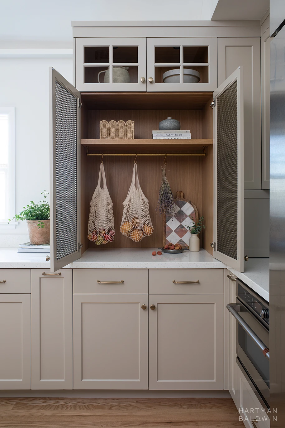 HartmanBaldwin Cambridge Residence California Ranch-Style Remodel Mesh Cabinet Open Custom tan-painted kitchen cabinetry with mesh-front doors open to showcase bags of fruit and drying lavender hanging from build-in brass rod