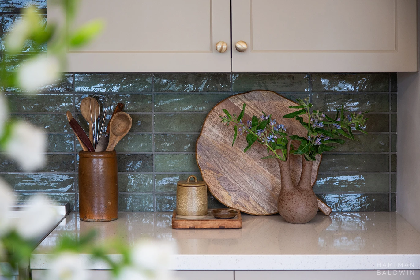 HartmanBaldwin Cambridge Residence California Ranch-Style Remodel Backsplash Detail Kitchen detail with tan-painted cabinetry, brass hardware, light stone countertops, blue-green tile backsplash, and wood and ceramic kitchen accessories