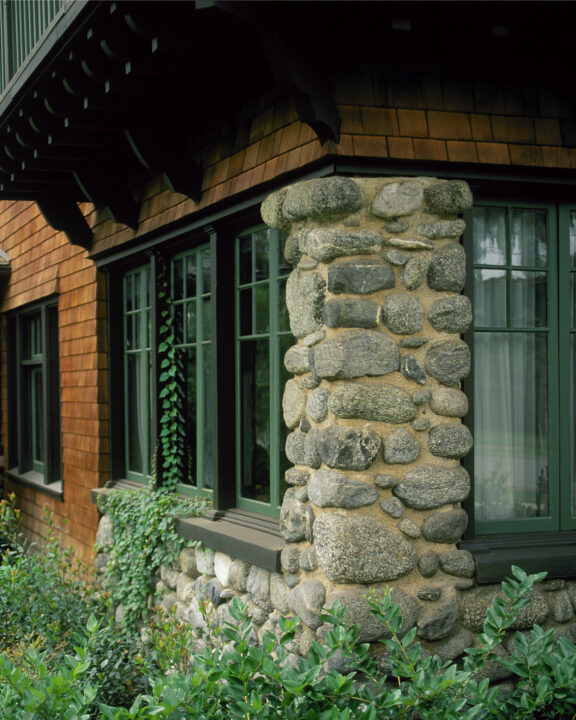 Craftsman-style home exterior details including stained-wood shingles, cobblestone pillar, divided light windows, and lush green landscaping