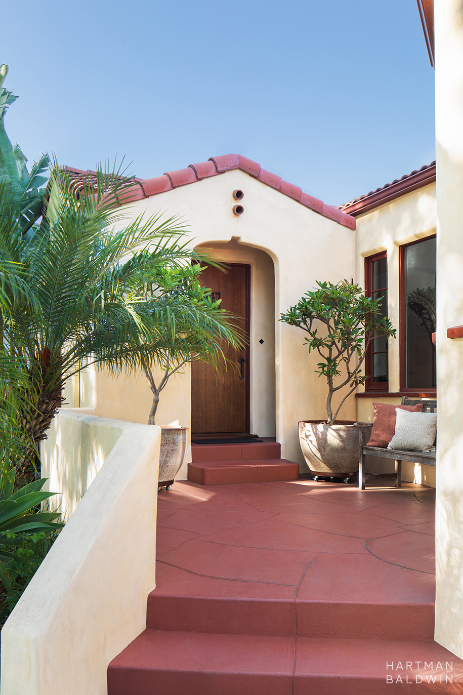 Spanish style home front porch with cream stucco walls, red flooring and clay tile roof, and wooden front door