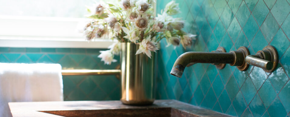 Bathroom sink with turquoise wall tiles, antique brass faucet, oxidized travertine sink, and floral arrangement