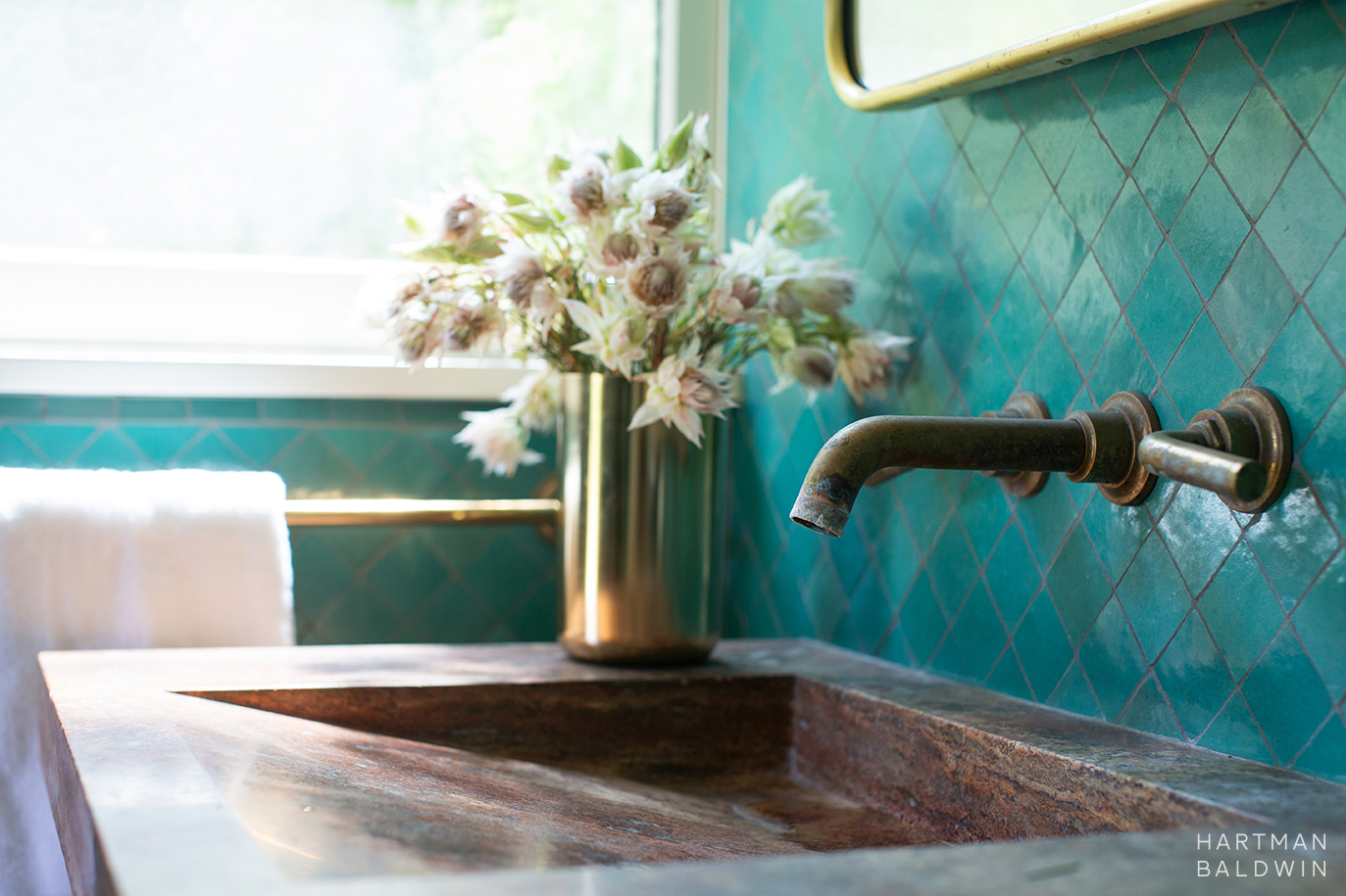 Bathroom sink with turquoise wall tiles, antique brass faucet, oxidized travertine sink, and floral arrangement