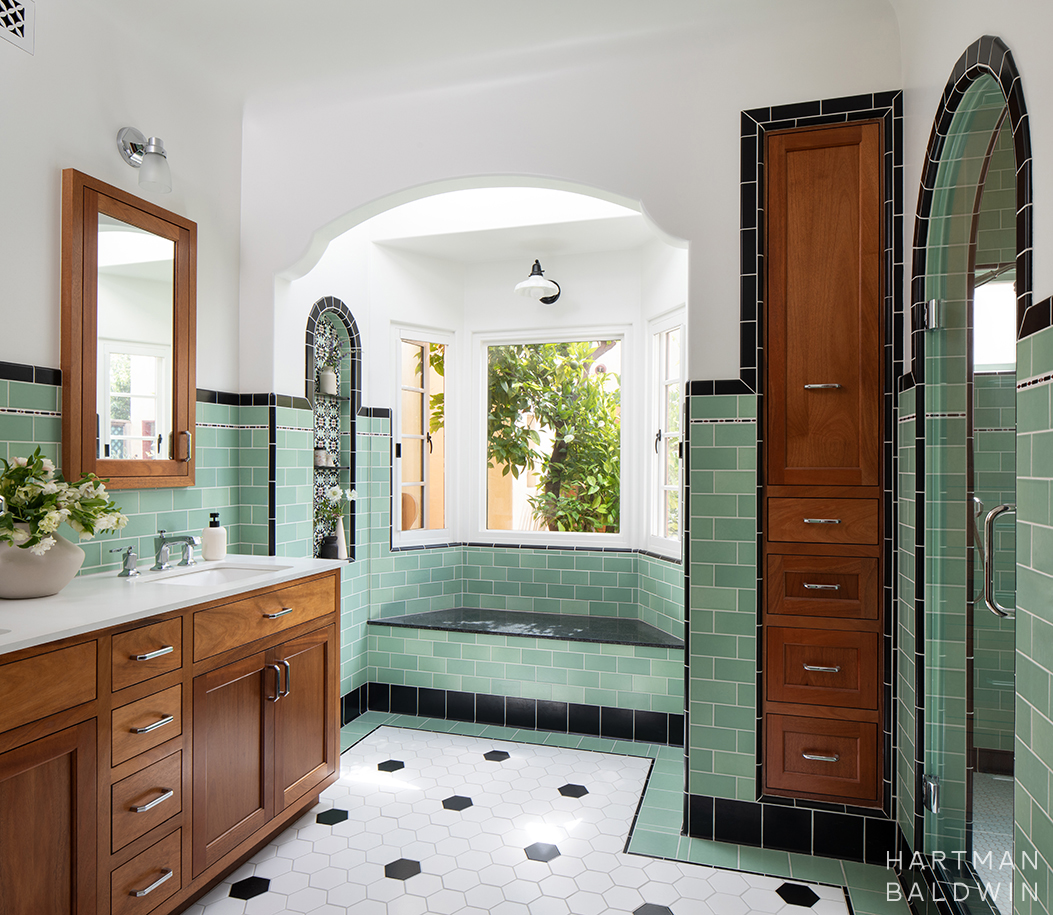 Spanish-style primary bathroom with warm mahogany cabinetry, seafoam-green wall tiles, decorative tiled niches, and a built-in window seat