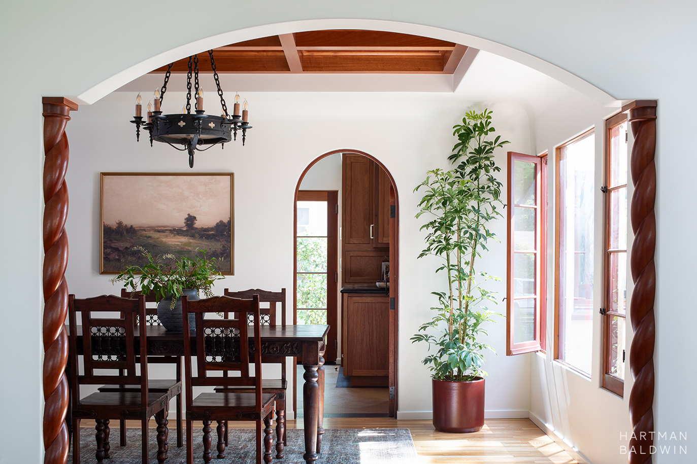 Remodeled Spanish-style dining room with dark-stained dining table, wrought iron chandelier, potted tree, and doorway to kitchen beyond seen through arched opening with barley-twist wood pillars