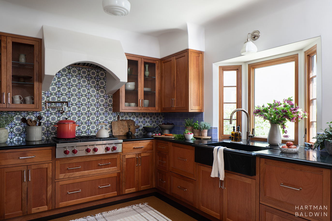 Remodeled Spanish-style kitchen with wood cabinetry, Sub Zero Wolf stove, arched plaster hood, black soapstone countertops, colorful patterned tile backsplash, and sink in front of a bay window