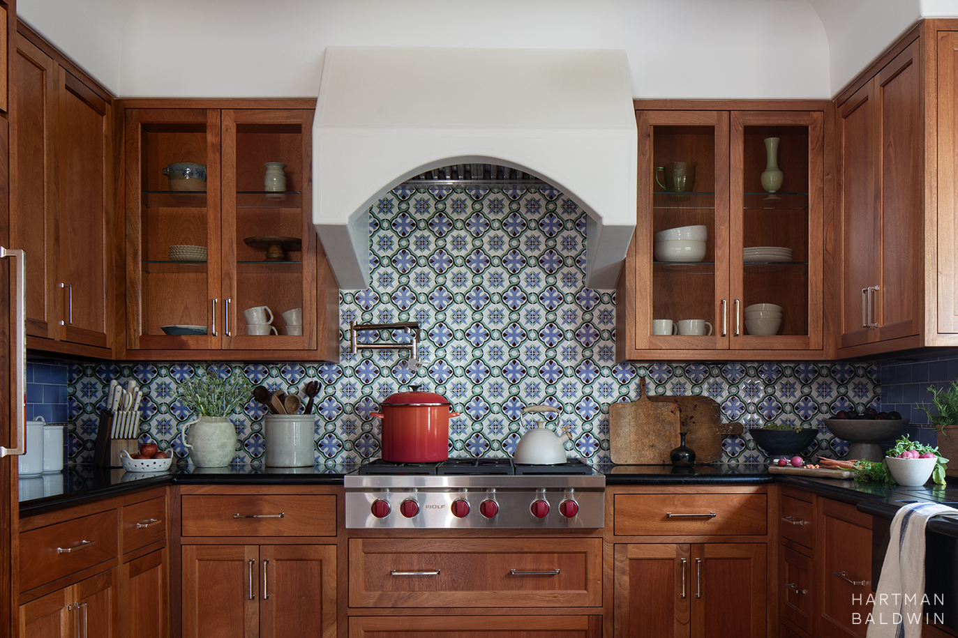 Remodeled Spanish-style kitchen with wood cabinetry, Sub Zero Wolf stove, arched plaster hood, black soapstone countertops, and a colorful patterned tile backsplash