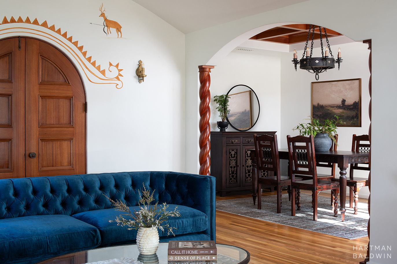 Remodeled Spanish-style living room and dining room with blue couch, dark-stained wood dining set, wrought iron chandelier, barley-twist pillars, and painted mural accents