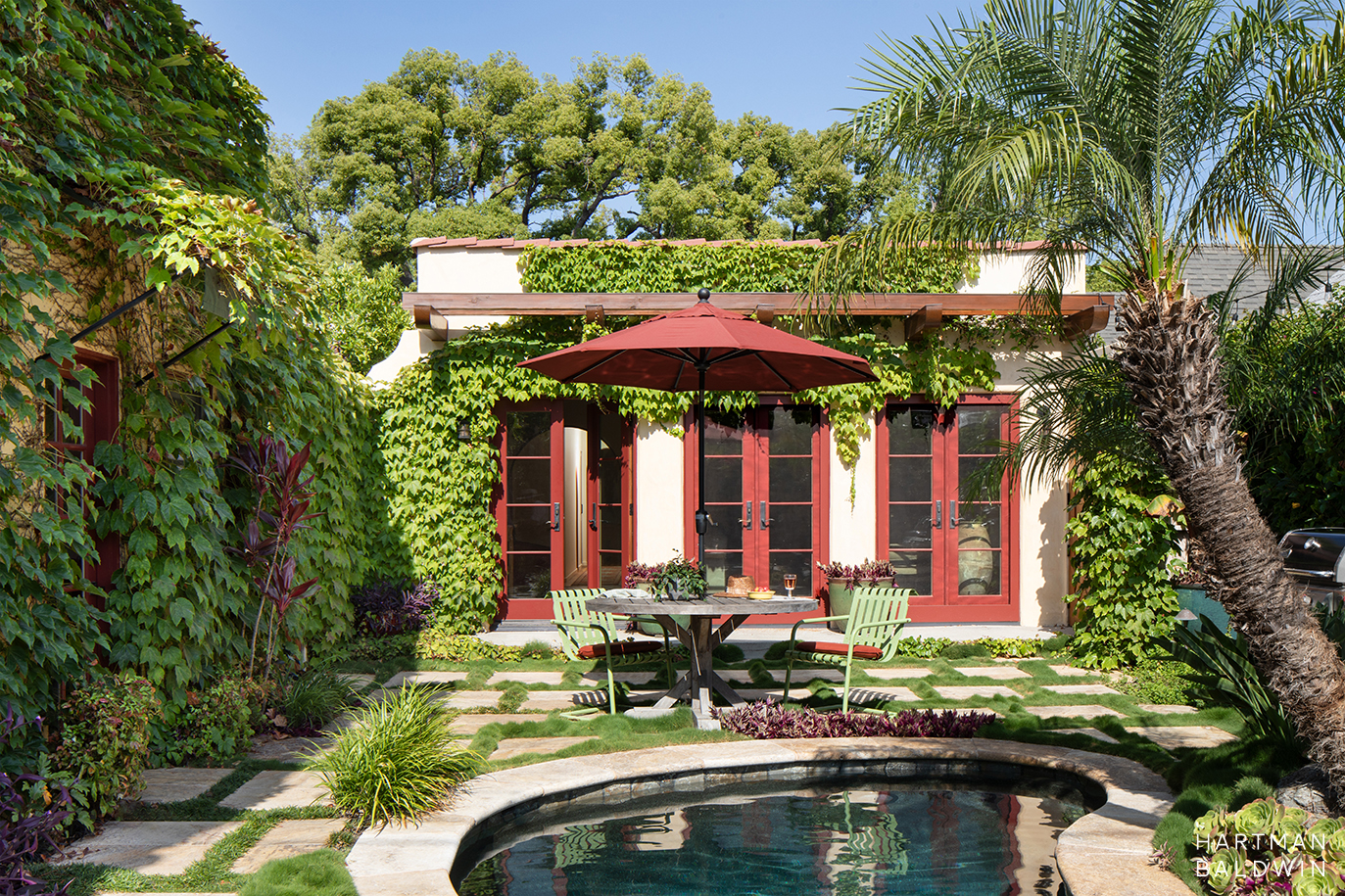 Spanish style backyard and home exterior with ivy covered cream stucco walls, red trim, wood trellis, patio dining table, and round pool