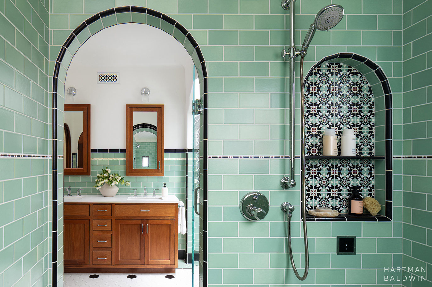 Blue-green tiled walk-in shower with arched niche featuring geometric accent tile and vanity seen through arched shower doorway