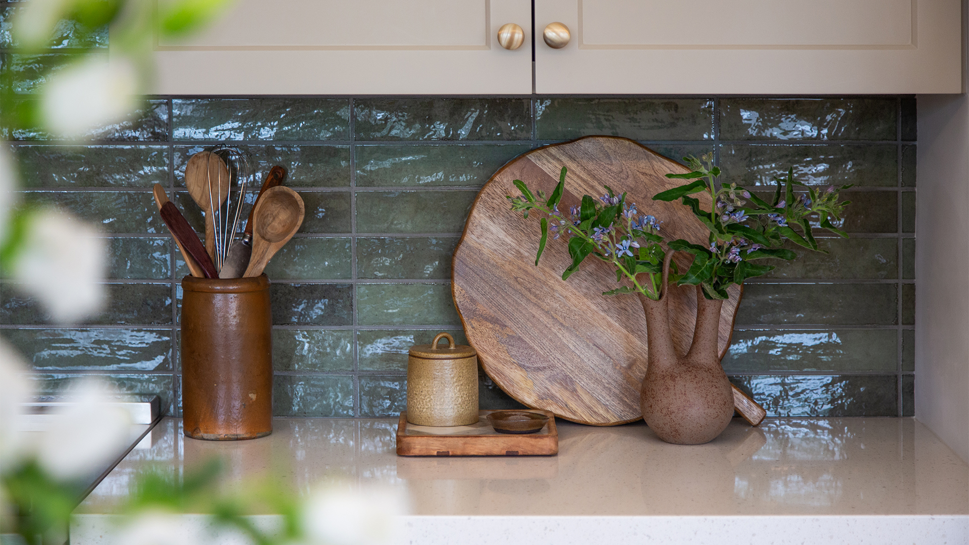 Kitchen detail with tan-painted cabinetry, brass hardware, light stone countertops, blue-green tile backsplash, and wood and ceramic kitchen accessories