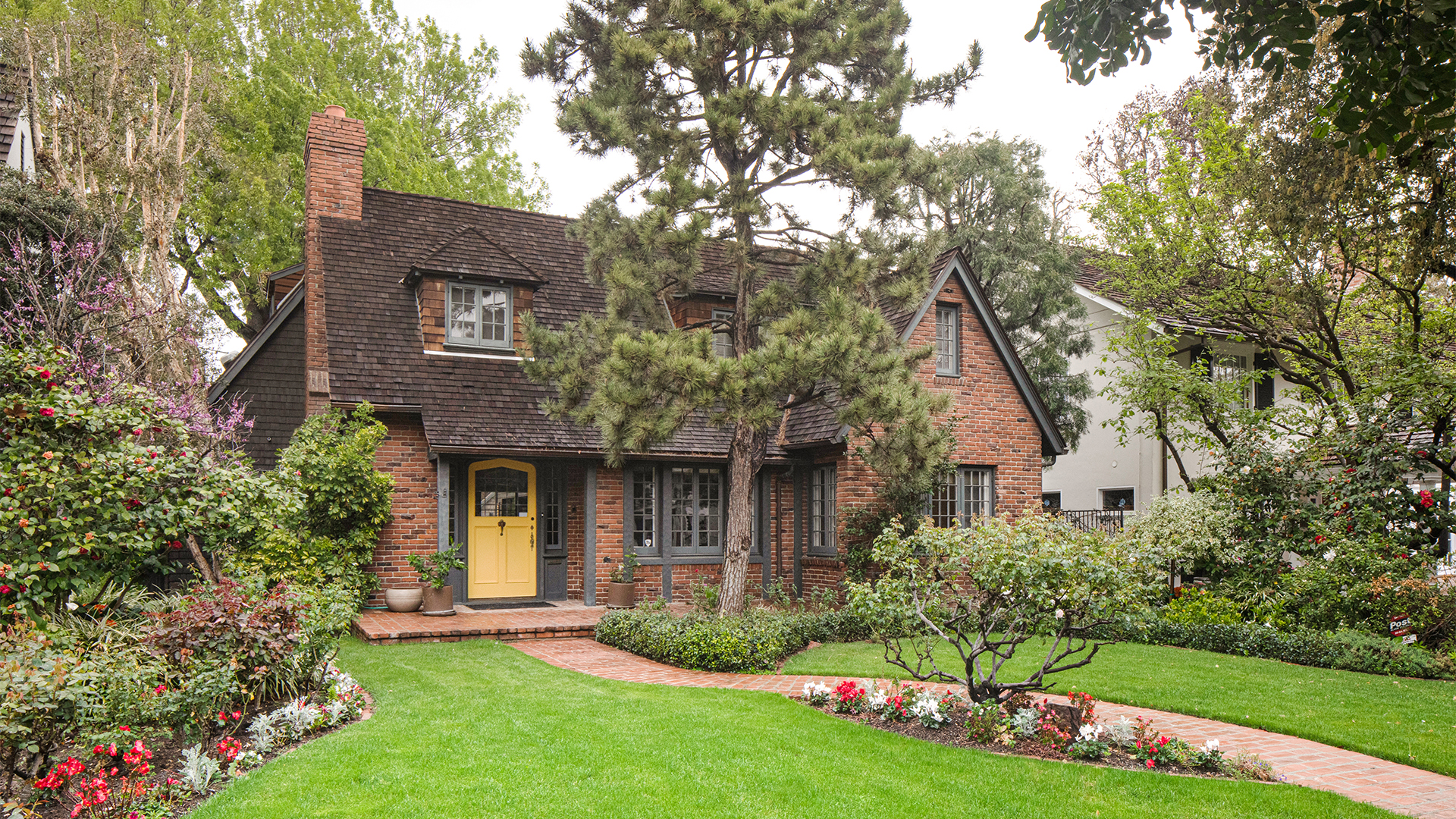 English cottage-style brick home exterior with yellow door, blue-grey trim, wood-shingle roof, and densely-planted front yard landscaping