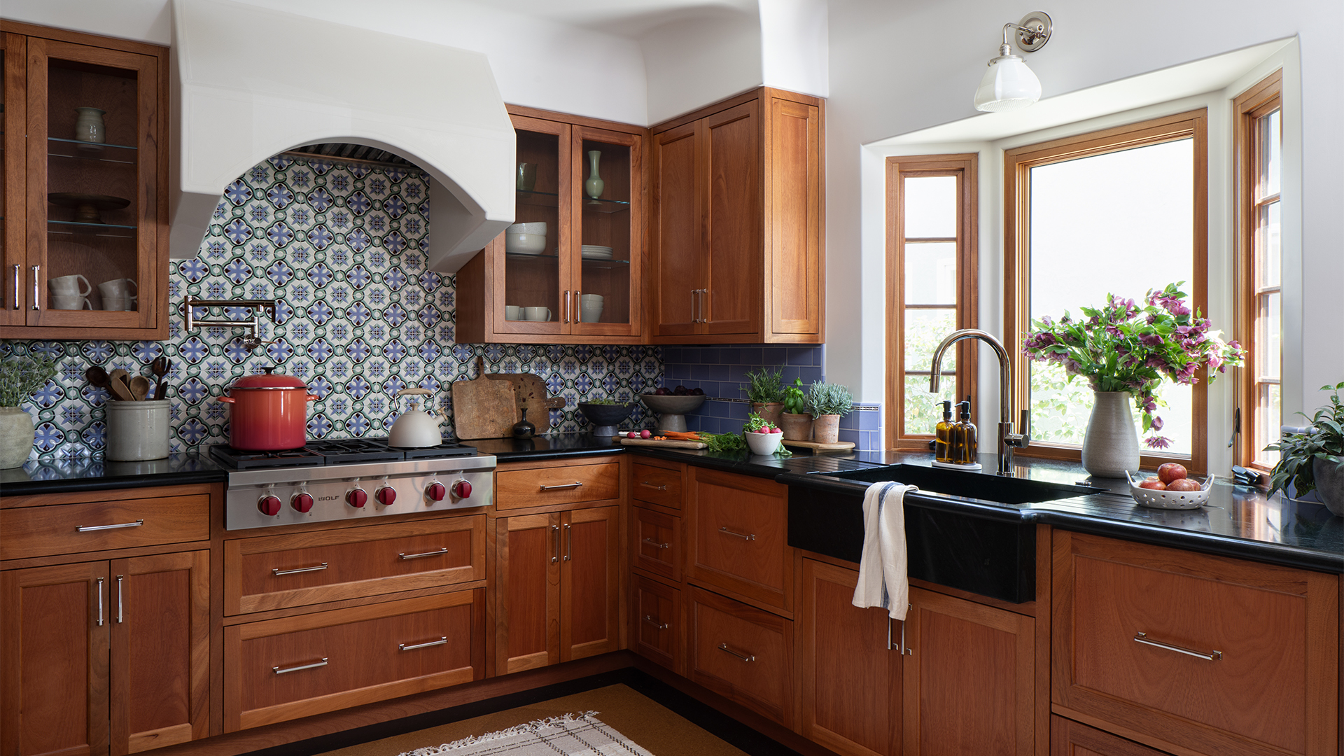 Remodeled Spanish-style kitchen with wood cabinetry, Sub Zero Wolf stove, arched plaster hood, black soapstone countertops, colorful patterned tile backsplash, and sink in front of a bay window
