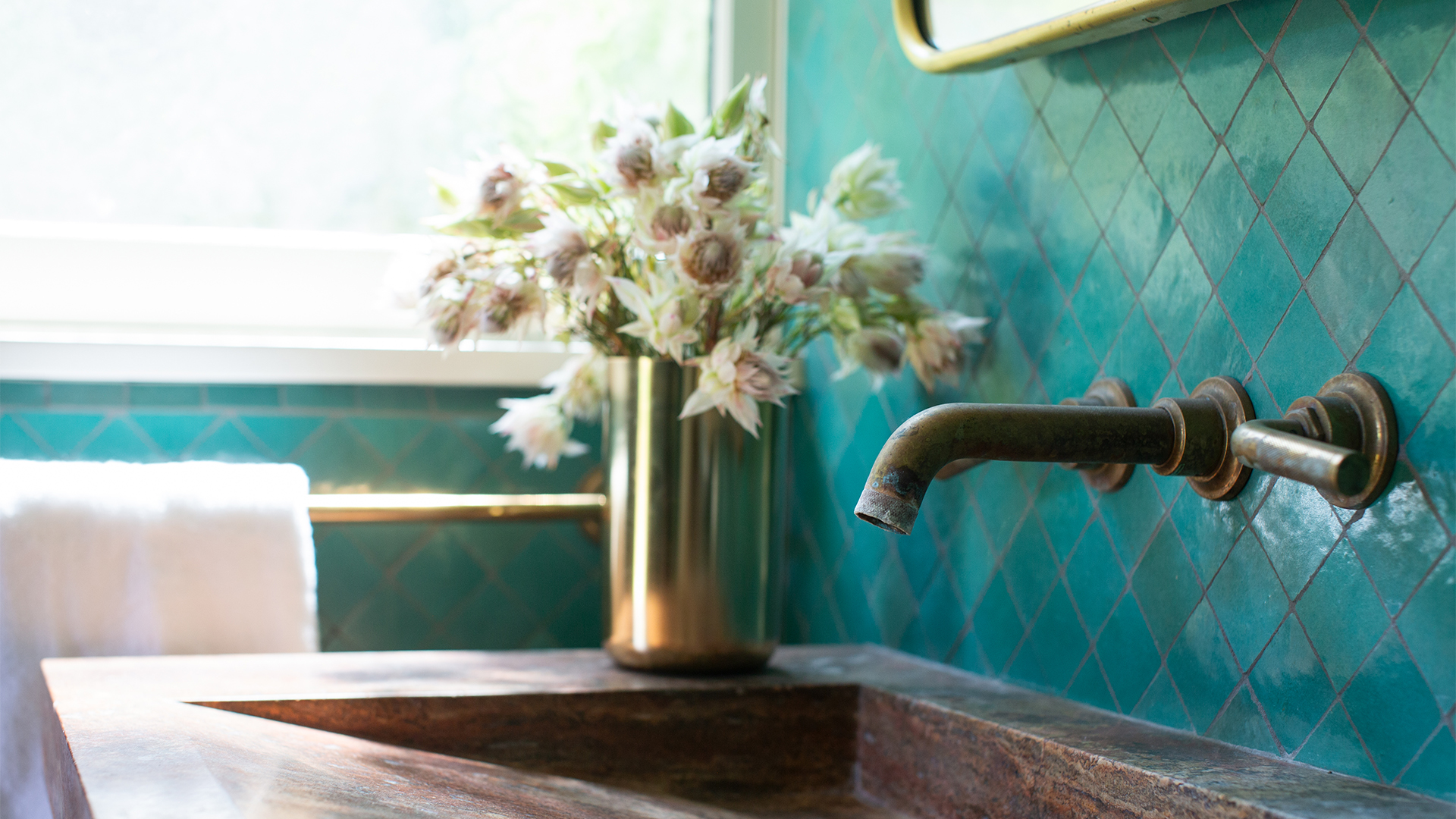 Bathroom sink with turquoise wall tiles, antique brass faucet, oxidized travertine sink, and floral arrangement