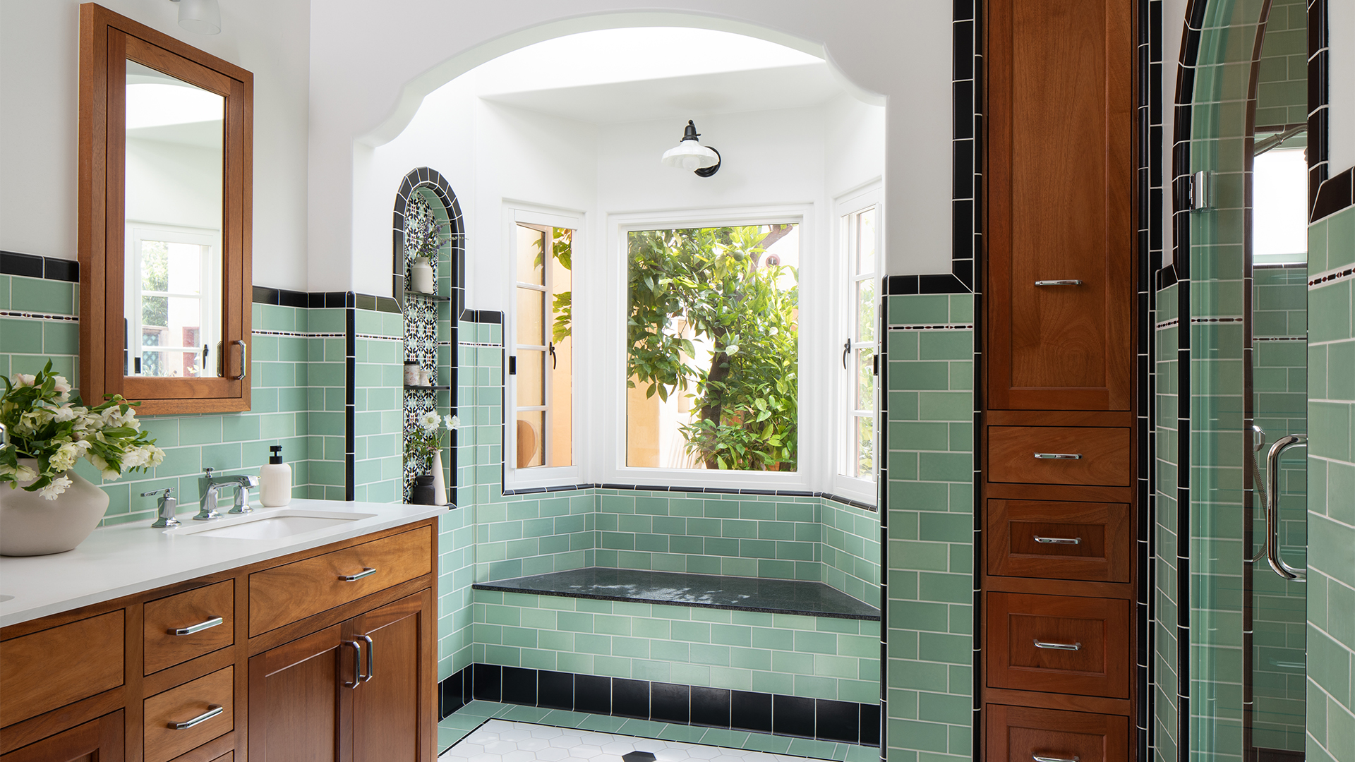 Spanish-style primary bathroom with warm mahogany cabinetry, seafoam-green wall tiles, decorative tiled niches, and a built-in window seat