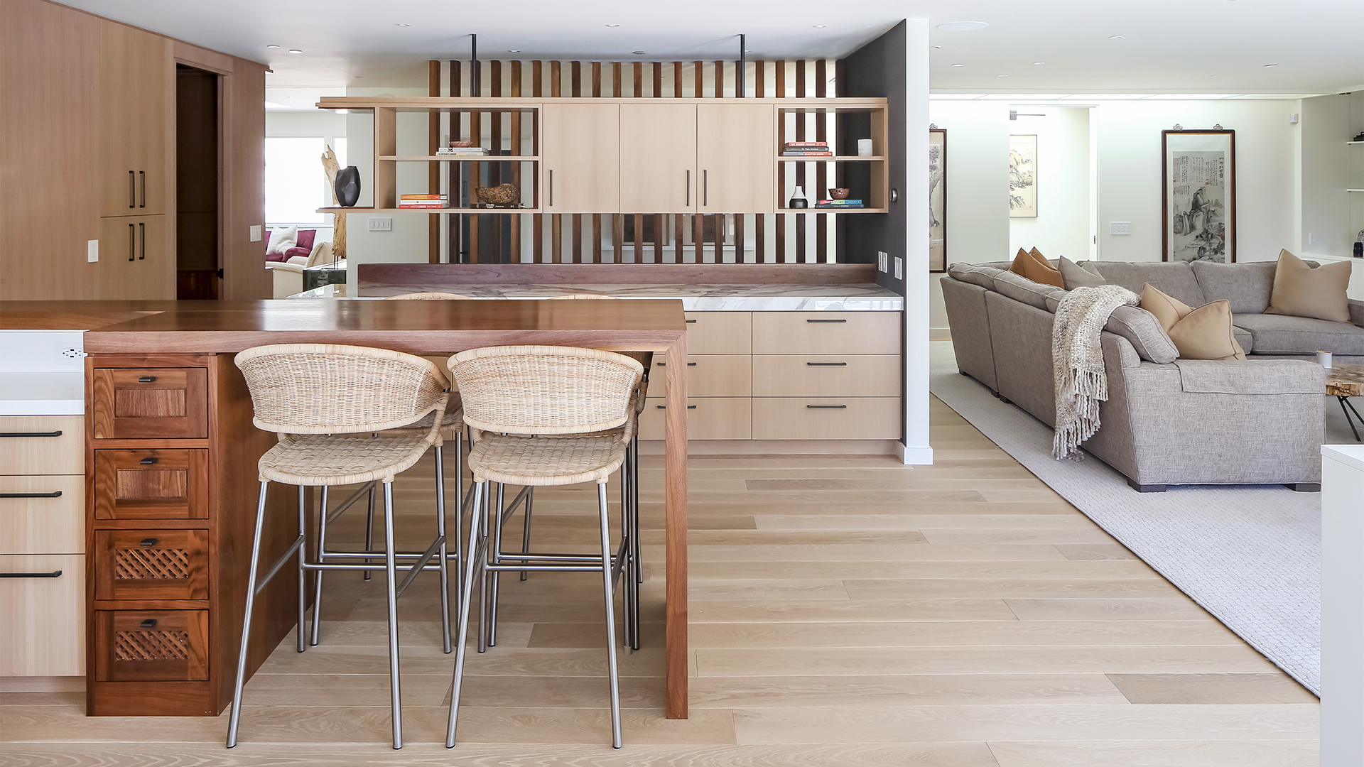 Waterfall-edge walnut island with bar seating, floating shelves, and quartzite buffet-style cabinet with dramatic multicolored veining. Living room with grey sectional couch and Chinese art pieces seen beyond.